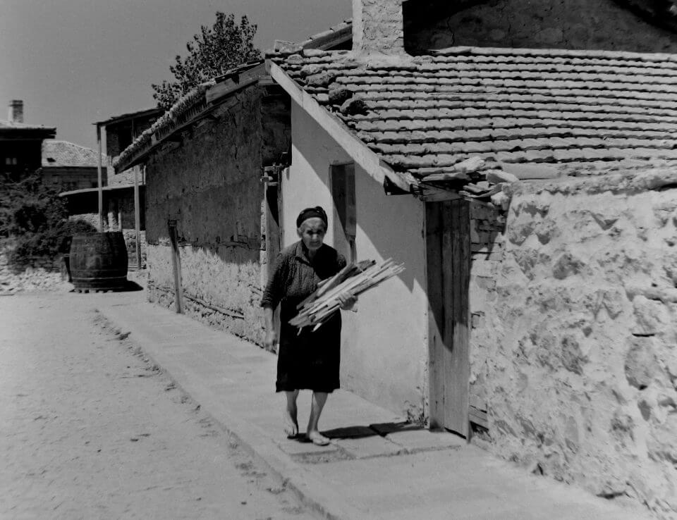 Staruszka z naręczem patyków przed chatą.
